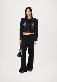 Veste noire courte avec des oiseaux brodés bleus et rouges, pantalon noir large et chaussures à imprimé léopard, assortis à un sac à main noir.