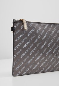 Pochette noire Versace Jeans avec imprimé logo argenté répété et tirette de fermeture éclair dorée sur une surface blanche.