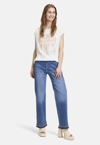 T-shirt blanc avec impressions graphiques, associé à un jean large bleu. Le jean présente des coutures décoratives au bas et est porté avec des sandales à plateforme beiges.