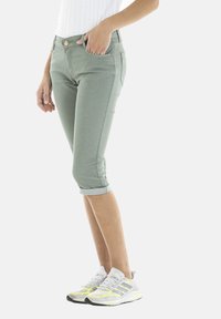 Jeans cropped vert menthe au design ajusté, avec des ourlets retroussés et un détail de bouton doré. Associés à des baskets blanches et grises avec des accents jaunes.