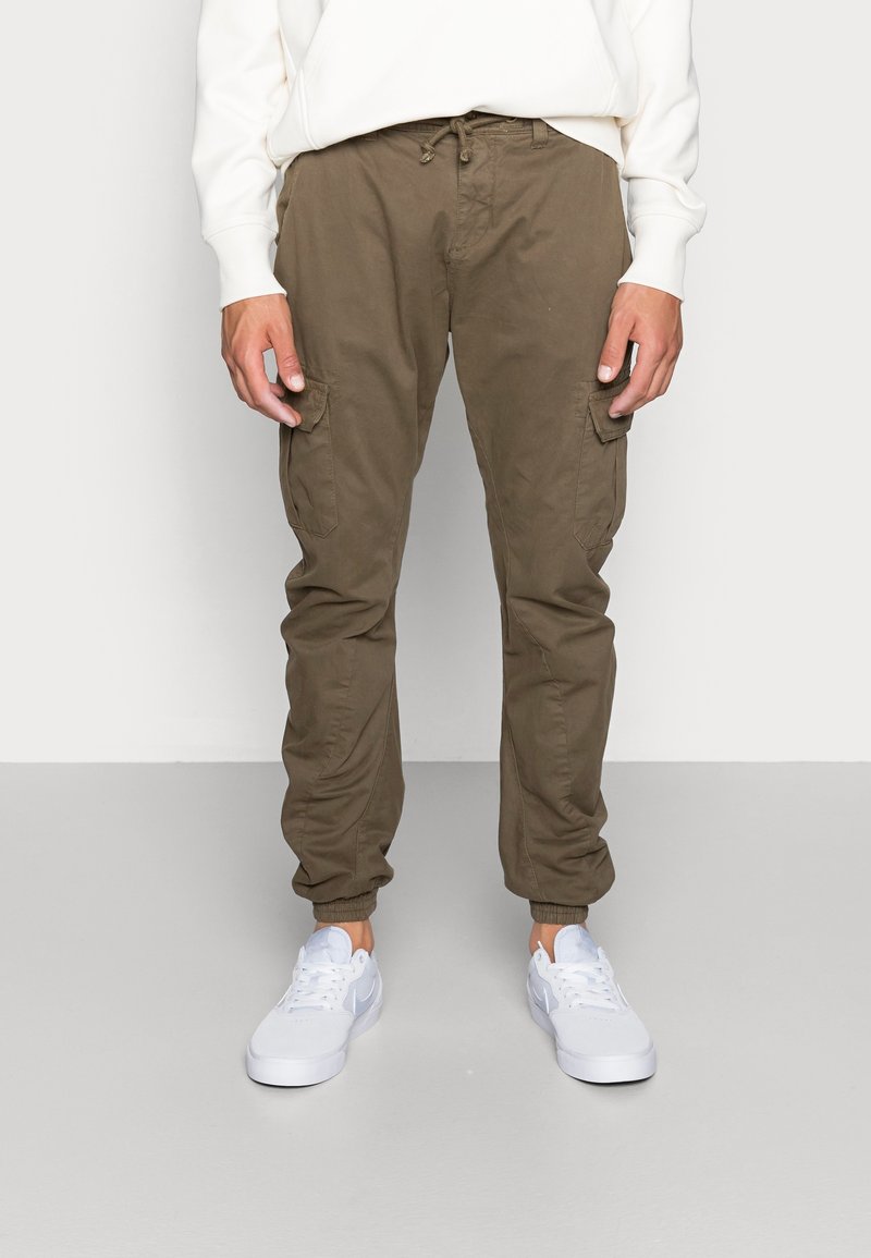 Homme portant un pantalon jogger cargo vert olive avec des poignets élastiques et des baskets blanches, debout devant un fond uni.