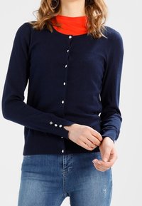 Navy blue cardigan made of soft fabric, featuring a round neckline, six button front closure, and ribbed cuffs. Worn over a red top.
