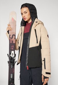 Beige und schwarze gepolsterte Skijacke mit Kapuze, Seitentaschen und Reißverschlüssen. Das Modell hält einen Ski mit Bindungen und steht vor einem neutralen Hintergrund.
