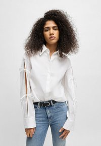 White button-up shirt with long sleeves featuring open side slits tied with ribbons. Worn with blue jeans and a black belt.