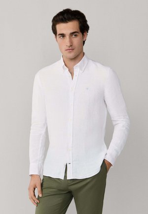 Hombre con camisa blanca de manga larga abotonada y pantalones verde oliva, de pie con una mano en el bolsillo contra un fondo liso.