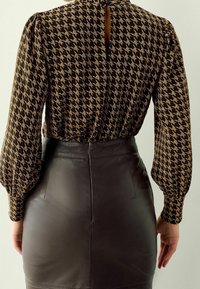 Blouse à manches longues noire avec un motif pied-de-poule, associée à une jupe en cuir noir ajustée. Détail avec ouverture en trou de serrure dans le dos de la blouse.