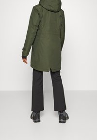 Personne debout, vêtue d'un parka à capuche vert olive, d'un pantalon noir et de chaussures de randonnée noires, vue de dos sur un fond uni.