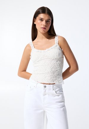 Top alb din dantelă cu bretele volan, design floral texturat, croi ajustat și tiv evazat, asortat cu blugi albi cu talie înaltă.