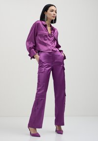 Lila Satin-Zweiteiler bestehend aus einem knöpfbaren Hemd mit Taschen und einer Cargo-Style-Hose. Glatte Textur, kombiniert mit spitzen Absätzen.