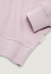 Sudadera de color púrpura claro con puños y dobladillo acanalados. Tejido suave, presenta una pequeña etiqueta de logo en el lateral. Textura suave y diseño sencillo.