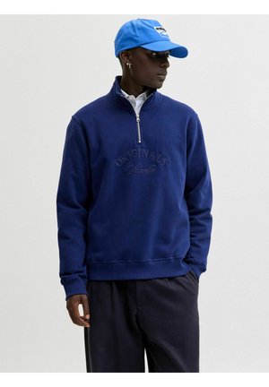 Tamno plava dukserica na zip s izvezenim natpisom "ORIGINALS" na prsima. Ima visoki ovratnik i rebraste manžete, u kombinaciji s plavom kapom.