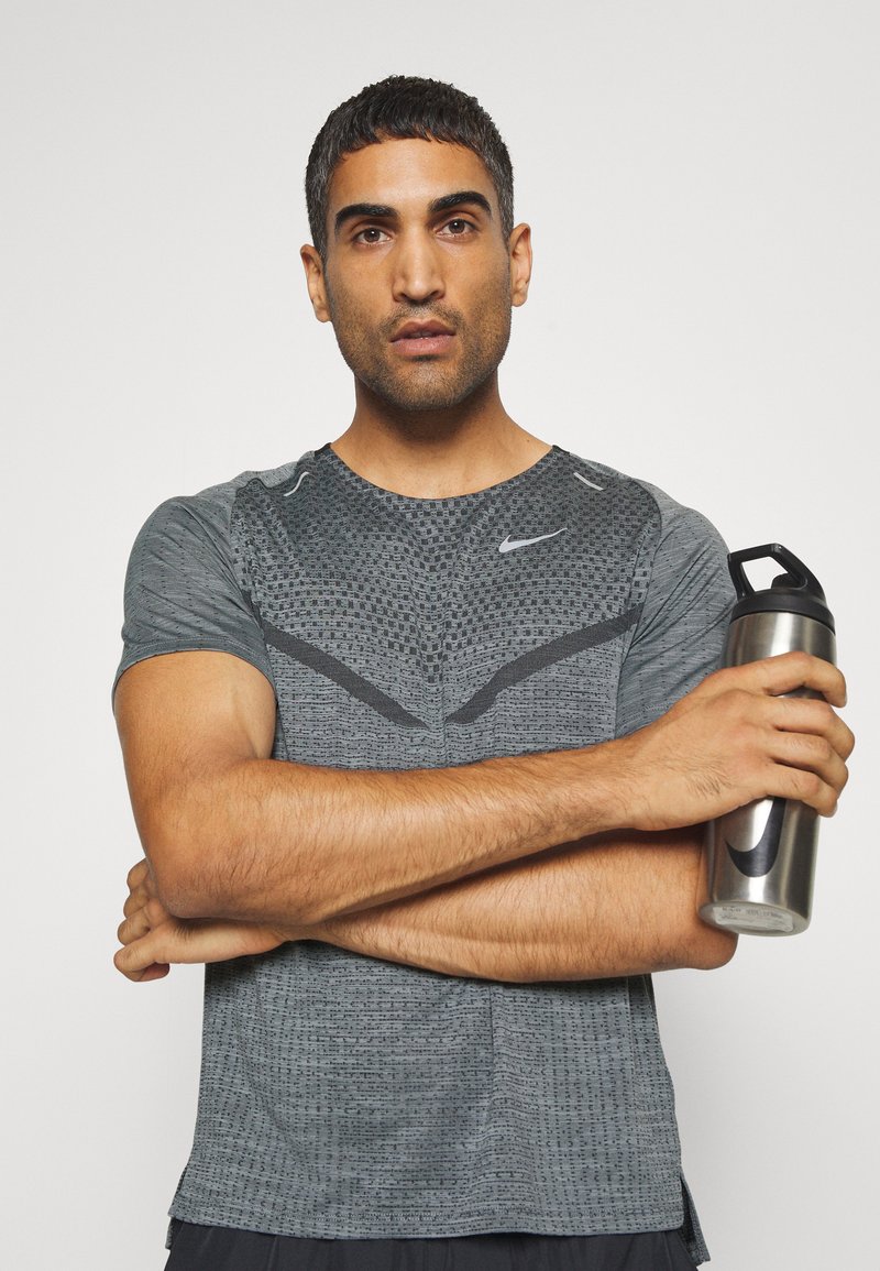 Nike Funktions-T-Shirt in Grau mit feuchtigkeitsableitendem Material, strukturiertem Muster, kurzen Ärmeln, runder Halslinie und schwarzen Akzenten, gehalten mit einer silbernen Wasserflasche.