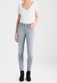 Jeans skinny gris claro con un aspecto desvanecido, que cuentan con un diseño de cinco bolsillos, un ligero desgaste y un cierre de botón. Combinados con una blusa blanca de escote en V.