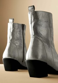 Botines metálicos plateados con acabado texturizado, cremalleras laterales y un tacón grueso negro. Cuenta con una lengüeta con asa para facilitar el calzado.