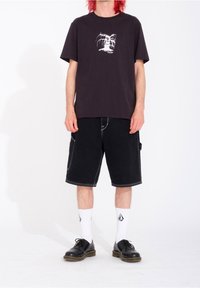 Svart bomullströja med vitt grafiskt tryck, kombinerad med svarta denimshorts, vita strumpor och svarta skor. Enkel, avslappnad design.