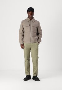 Bruine zip-up jas met twee borstzakken, gecombineerd met lichtgroene cargobroeken en zwarte chunky schoenen. Het model draagt een zwarte pet.