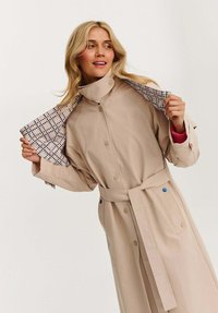Žena v béžovém trenčkotě s páskem, který odhaluje vzorovanou podšívku, se usmívá a dívá se do strany na neutrálním pozadí.