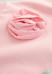 Roza majica z detajlom iz teksturiranega materiala v obliki cvetov, s softness in mehkimi gubami. Tkanina izgleda gladko in lahko, z diskretnim rebrastim vzorcem.