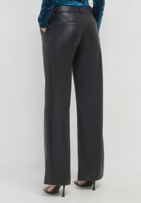 Donna che indossa pantaloni in pelle neri a gamba larga, una maglietta in velluto blu e sandali neri con tacco alto, in piedi con le mani nelle tasche.