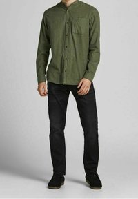 Man i olivgrön skjorta med knäppning och mandarinkrage, svarta jeans och svarta skor, står mot en grå bakgrund.