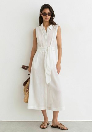 Vrouw draagt een mouwloze witte midi-jurk met knopen en een tailleceintuur, zonnebril, sandalen en houdt een strooien handtas vast tegen een witte muur.