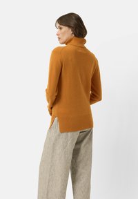 Svetr s rolákem v spálené oranžové barvě, vyrobený z měkké látky. Má žebrované manžety a lem s bočními rozparky pro snadný pohyb.