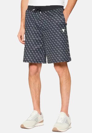 Homme portant un short noir à motifs avec des poches latérales et des baskets blanches, debout les mains dans les poches sur fond blanc.