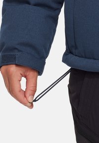 Blå tygjacka med strukturerad yta, som har en hand som drar i en justerbar snodd vid nederkant, tillsammans med en del av svarta byxor.