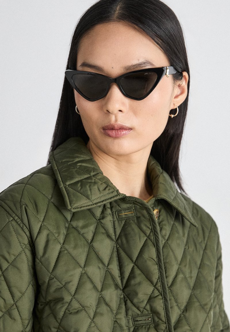 Crne sunčane naočale u obliku mačjih očiju s kutnim dizajnom, nosene uz jaknu od maslinasto zelene tkanine s quilted uzorkom, koja ima okovratnik i dijamantni uzorak.