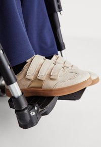 Pieds portant des baskets beige à scratch reposant sur le repose-pieds d'un fauteuil roulant, personne vêtue d'un pantalon bleu marine.
