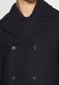 Dobbeltbrystet mørkeblå frakke med store revers, tekstureret uldstof og fire sorte knapper. Har en struktureret pasform og rene linjer.