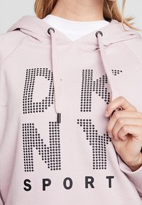 Sweat à capuche rose clair avec texte noir "DKNY SPORT", orné de clous métalliques. Comprend une capuche avec cordon de serrage et une texture de tissu doux.