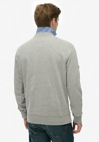 Hombre con sudadera gris clara sobre camisa de cuello azul y jeans oscuros, mostrado de espaldas contra un fondo blanco.