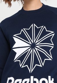 Námorně modrá mikina s velkým bílým geometrickým designem výbuchu hvězdy na hrudi, s žebrovanými manžetami a límcem.