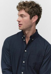 Jonge man met krullend bruin haar, gekleed in een donkerblauwe bloes met knopen, die naar links kijkt tegen een eenvoudige lichte achtergrond.