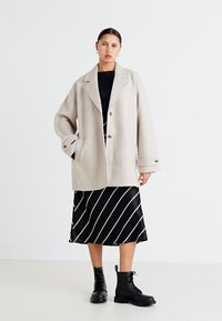 Beige overcoat dengan lapel notch, tiga kancing depan, dipadukan dengan atasan hitam dan rok midi bergaris hitam-putih, diselesaikan dengan sepatu bot hitam.