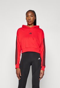 Rode cropped hoodie met een logo aan de voorkant, een trekkoord-hoodie en mouwen met zwarte strepen, gecombineerd met zwarte leggingen met een logo op de bovenbeen.