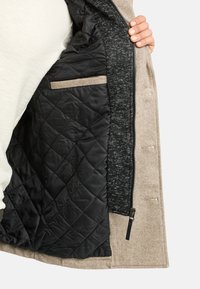 Esterno beige con interno trapuntato nero; presenta una fodera grigia testurizzata e una cerniera. Include una tasca interna con rifinitura a contrasto.