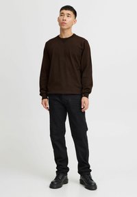 Camisa marrón de manga larga con cuello redondo, combinada con pantalones de mezclilla negros y botas negras con cordones. Diseño simple, silueta ajustada.