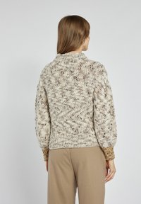 Gruby sweter w kolorze off-white z brązowymi drobinkami, z wysokim kołnierzem, teksturowanymi rękawami i ściągaczem na dole. Nosi się go na beżowych spodniach.