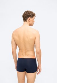 Άνδρας, που φαίνεται από πίσω, φοράει σκούρο μπλε μπόξερ με πορτοκαλί ζώνη μέσης με την ένδειξη "Lacoste" σε απλό φωτεινό φόντο.