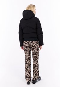 Persona de espaldas con una chaqueta acolchada con capucha negra, pantalones de estampado de leopardo y botas de esquí negras, frente a un fondo blanco.