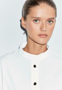 Witte overhemd met een knoopsluiting aan de voorkant met zwarte knopen en een gladde, textuurgevel. Effen achtergrond benadrukt het ontwerp van het shirt.