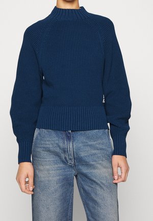 Marineblå ribstrikket sweater med høj krave og lange puffede ærmer, båret med lyseblå jeans.