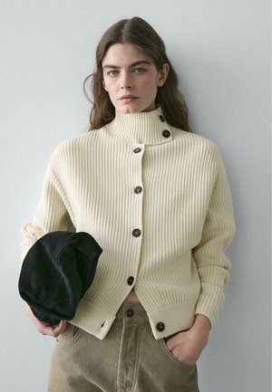 Donna che indossa un maglione crema a coste con bottoni e tiene un berretto nero, con una mano in tasca, su sfondo semplice.