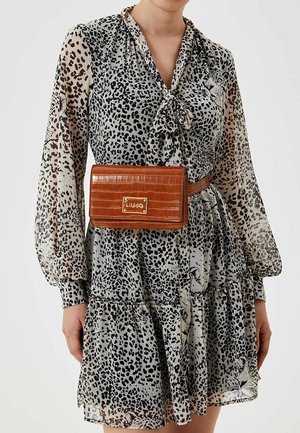 Kobieta ubrana w przezroczystą sukienkę w lamparcie cętki z długimi rękawami i zawiązywanym dekoltem, uzupełnioną brązową torebką na pasku o teksturze krokodyla.