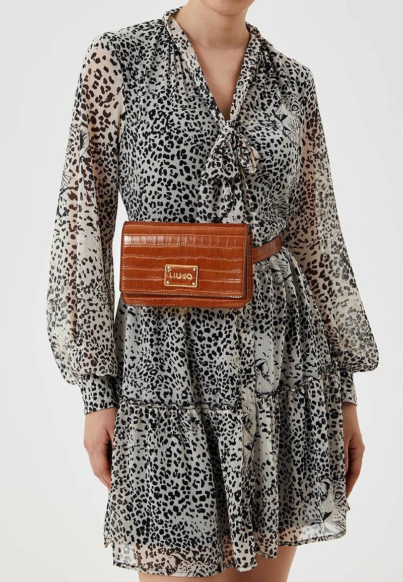 Kvinde iført en gennemsigtig leopardprintkjole med lange ærmer og bundet halsudskæring, accessoriseret med en brun bælteväske med krokodilletekstur.