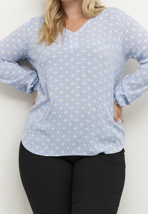 Vrouw met een lichtblauwe, met patronen bedrukte blouse met lange mouwen en V-hals en een zwarte broek, handen op de heupen tegen een witte achtergrond.
