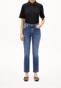 Person wearing a black short-sleeve polo shirt, high-waisted blue jeans, and black high-heeled sandals, standing with one hand in pocket.