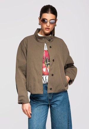 Frau trägt braune Jacke mit Knöpfen über einem grafischen T-Shirt, blaue Jeans, Sonnenbrille und goldene Creolen, posiert mit einer Hand in der Tasche.